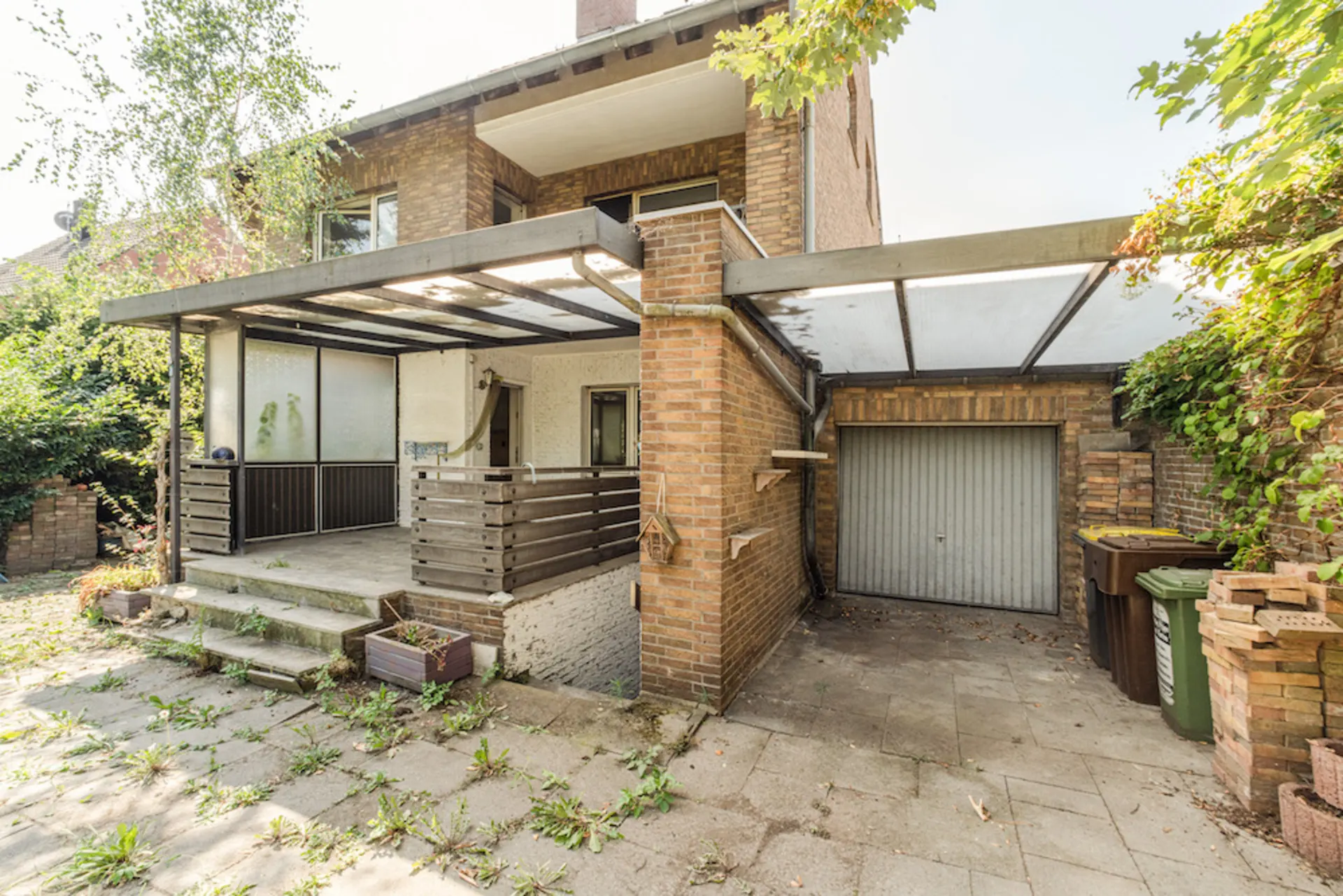 Backsteinwohnhaus mit überdachter Terrasse und Garage, umgeben von Pflanzen und Mülltonnen.