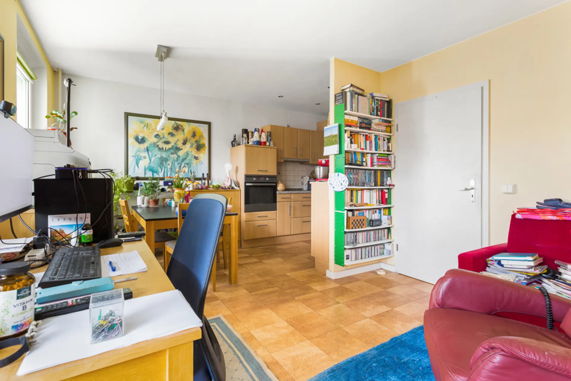 Wohnzimmer mit Schreibtisch, Bücherregal, rotem Sessel und offener Holzküche im Hintergrund.