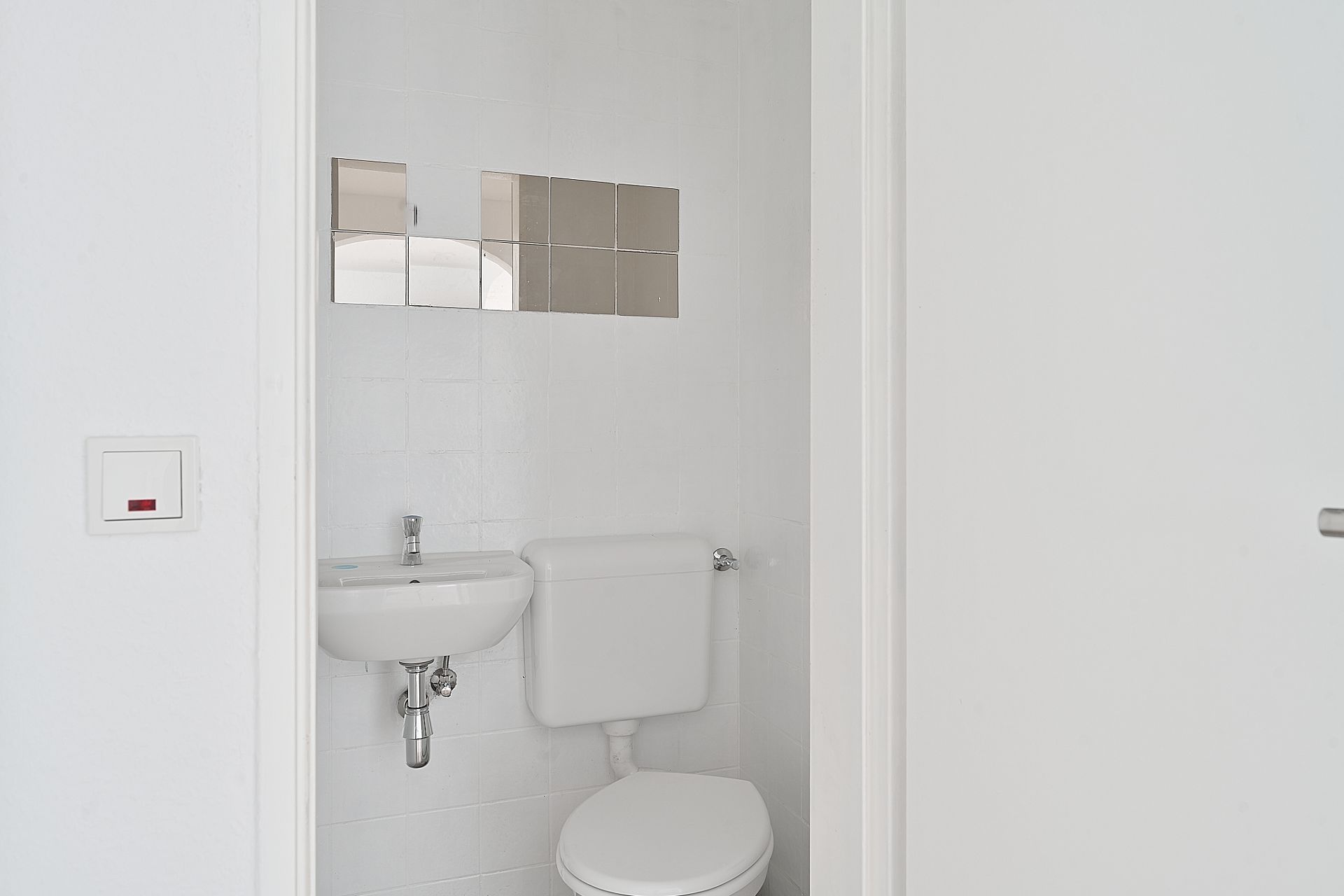 Kleines, weiß gefliestes Bad mit Toilette, kleinem Waschbecken und Spiegelkacheln an der Wand.