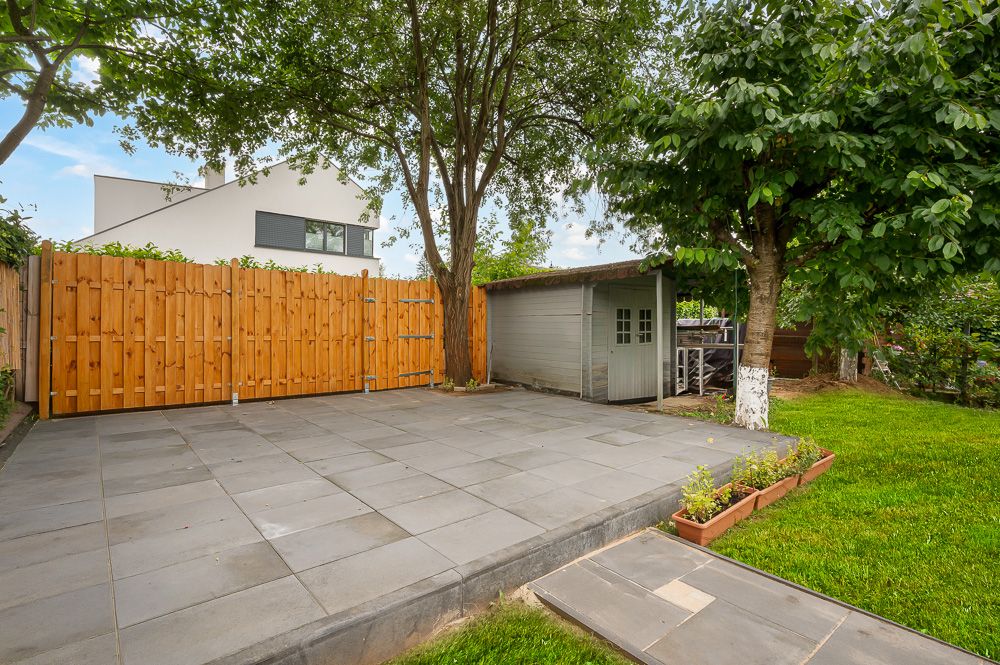 Gepflasterte Terrasse mit Gartenhaus, Holzlattenzaun und Bäumen im Garten.