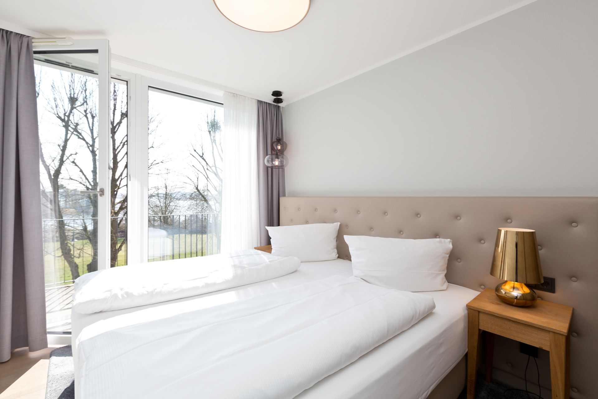 Helles Schlafzimmer mit Doppelbett, Nachttisch und grossem Fenster mit Blick nach draußen.