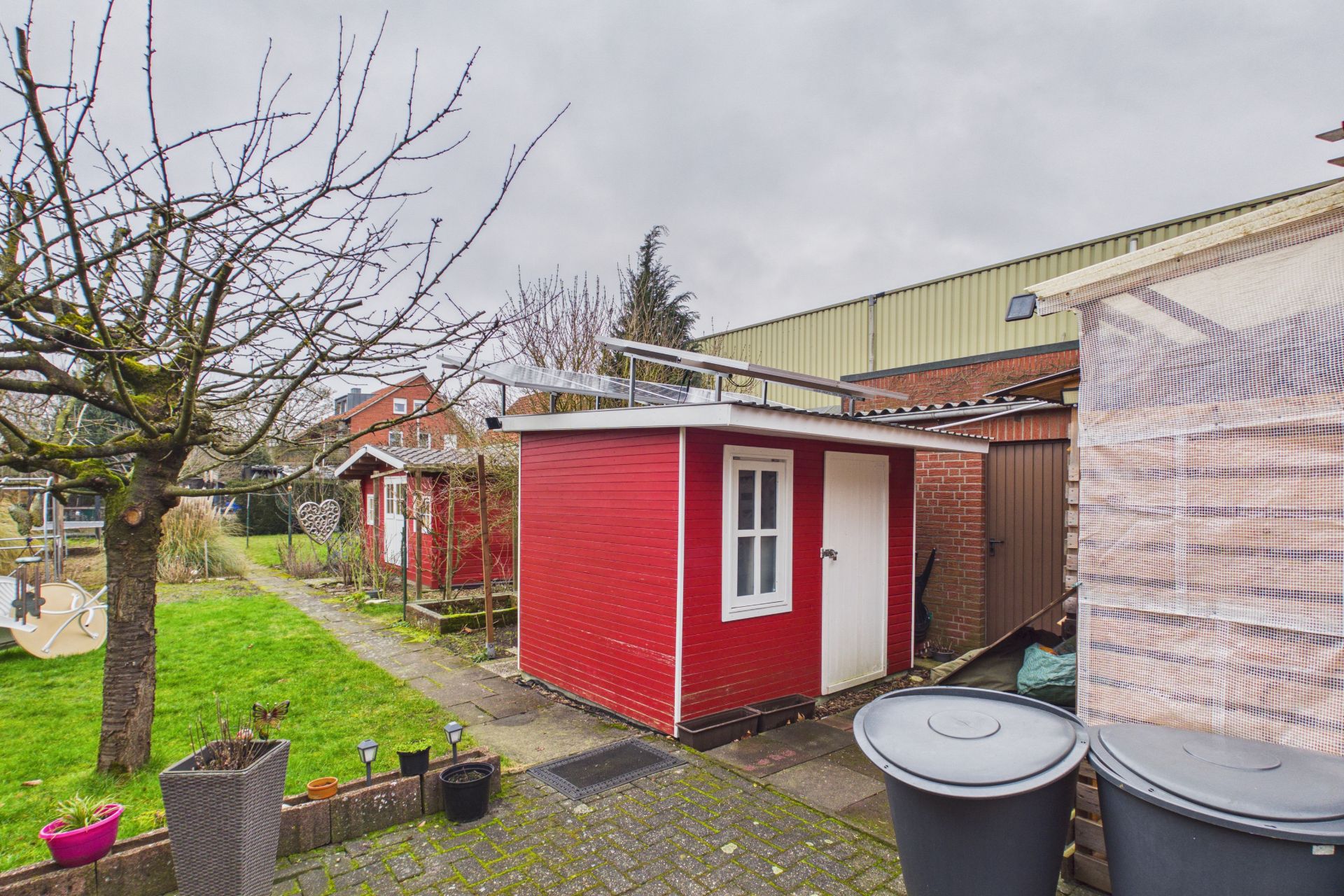 Garten mit rotem Geräteschuppen, zwei Mülltonnen und mehreren Bäumen bei bewölktem Himmel.