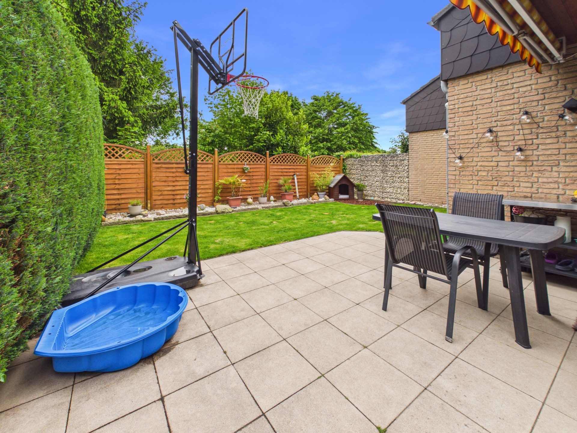 Gepflasterte Terrasse mit Tisch, Stühlen, Basketballkorb und Sandmuschel im Garten.