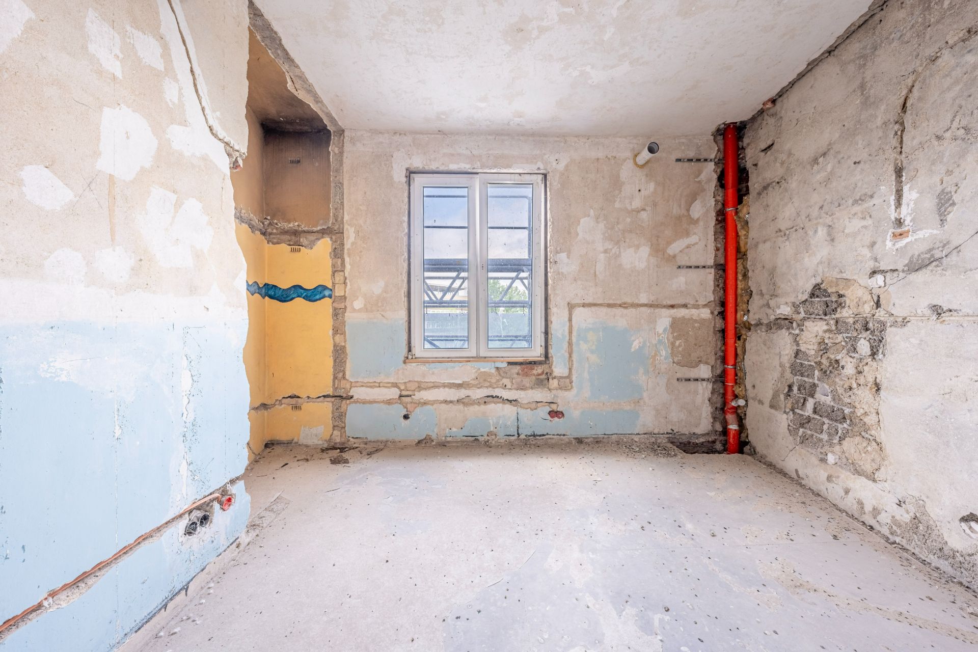 Leerer Raum mit abgenutzten Wänden, einem Fenster und sichtbaren Rohren.