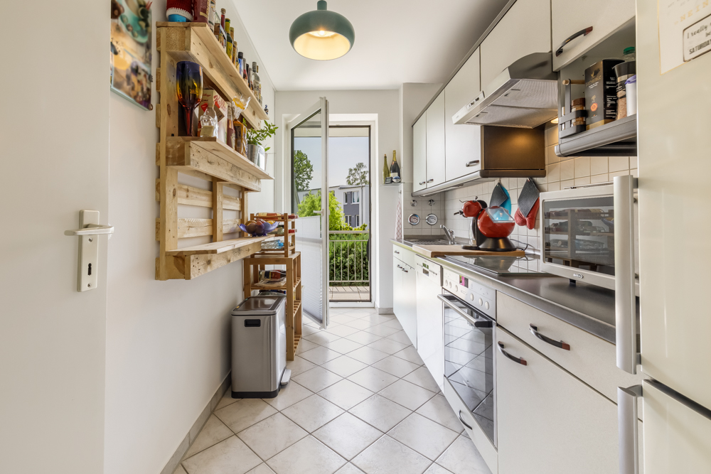 Helle, schmale Küche mit weißen Schränken, Regalen und Glastür zum Balkon.