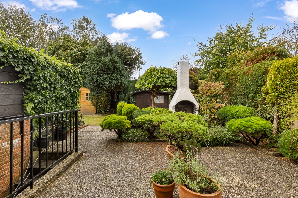 Gepflegter Garten mit Terrasse, Grillkamin, Sträuchern und zwei kleinen Gartenhäusern.