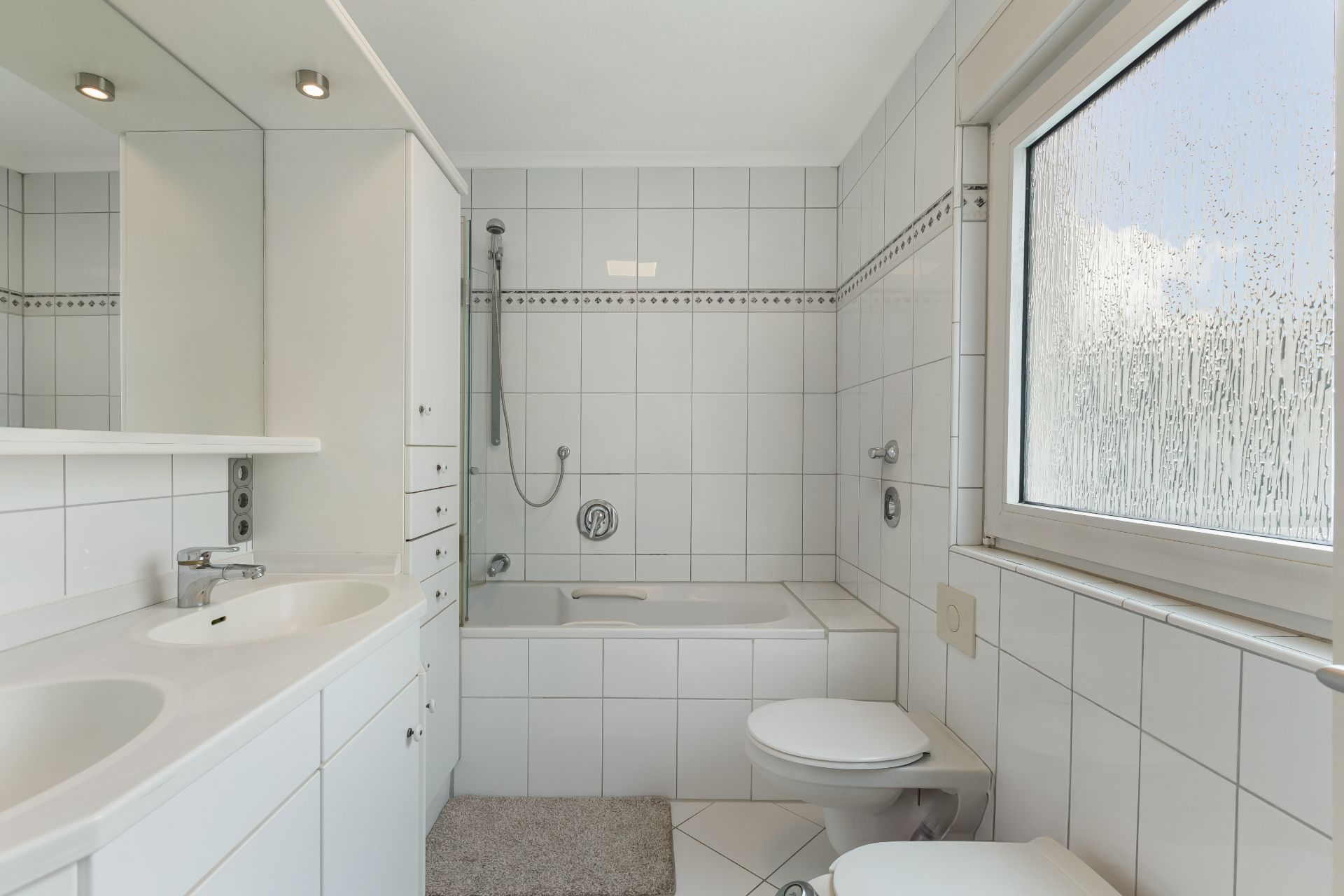 Helles Badezimmer mit Doppelwaschbecken, Badewanne, Toilette und Fenster.