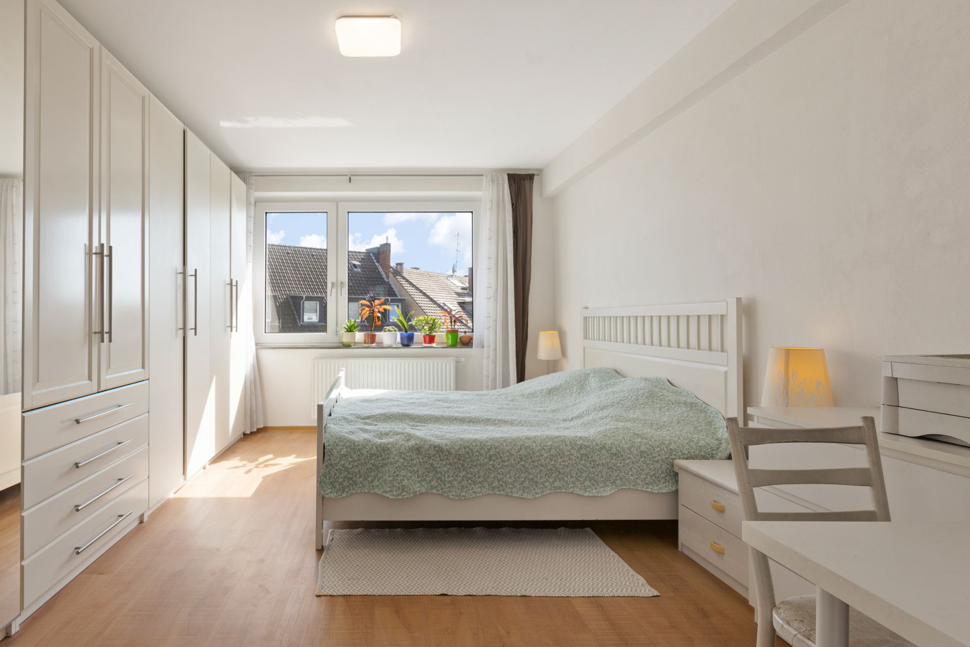 Helles Schlafzimmer mit Bett, weißen Schränken, Schreibtisch und großen Fenstern mit Topfpflanzen.