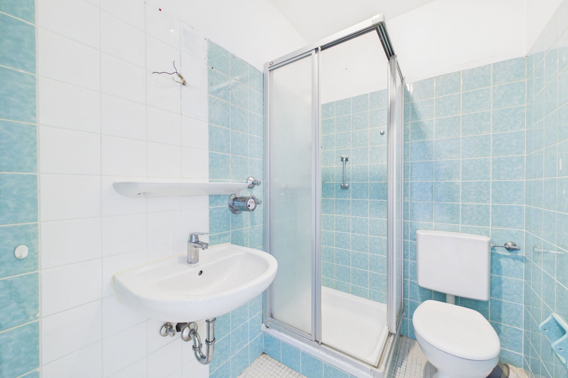 Kleines Badezimmer mit Dusche, Waschbecken und Toilette, hellblaue Fliesen an den Wänden.