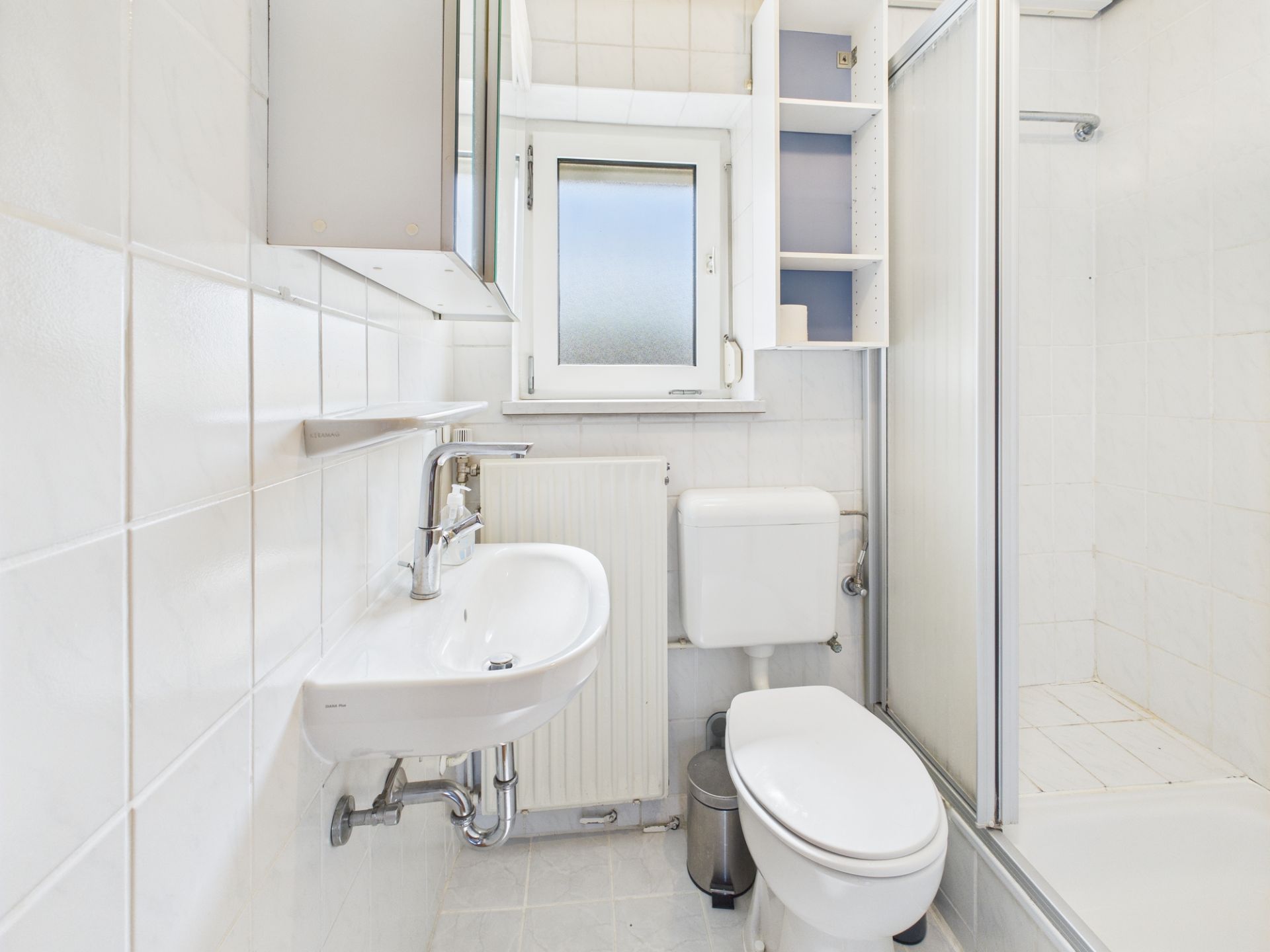 Kleines Bad mit Waschbecken, Toilette, Dusche, Spiegel und Fenster.