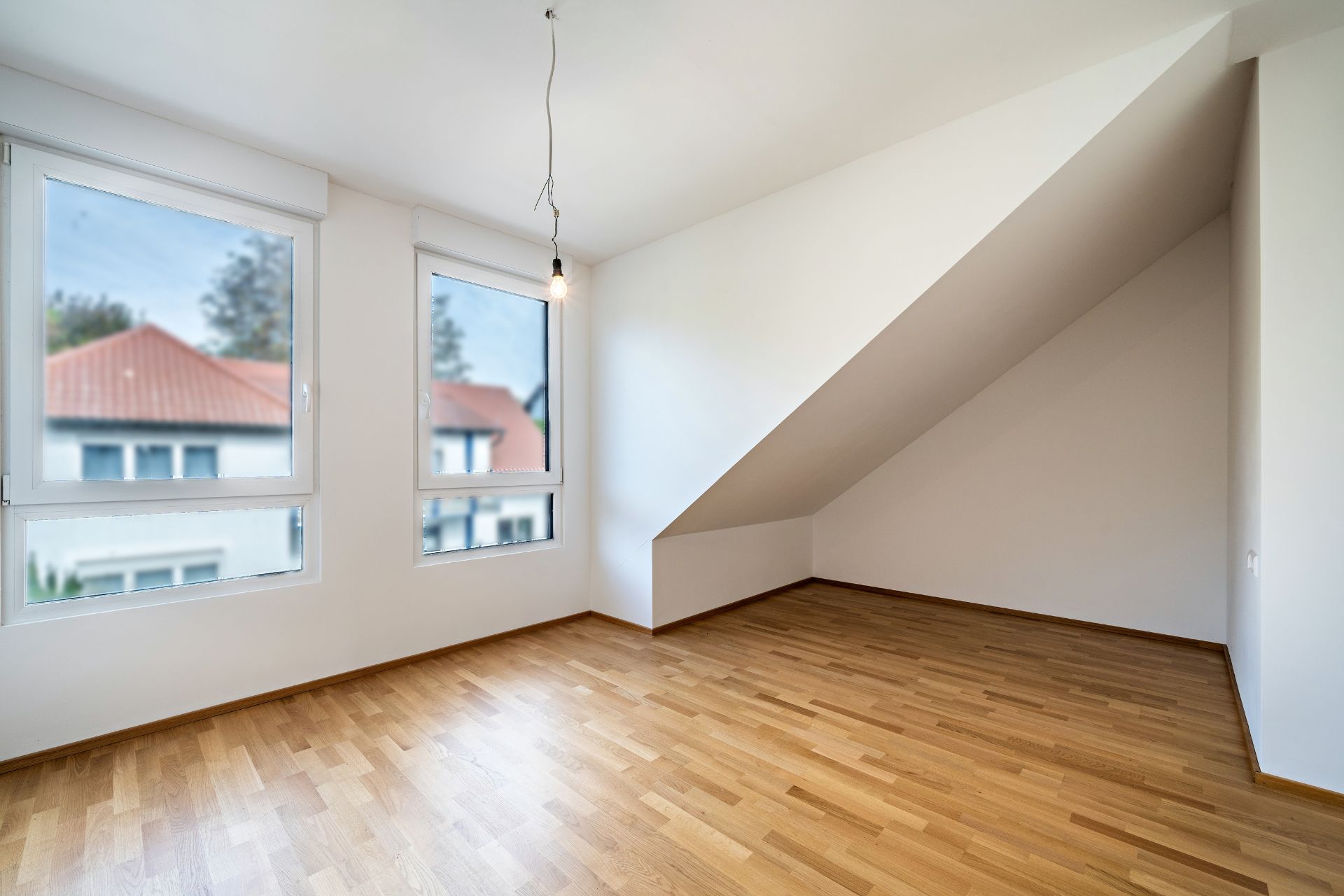 Leeres Zimmer mit Parkettboden, zwei Fenstern und Dachschräge.