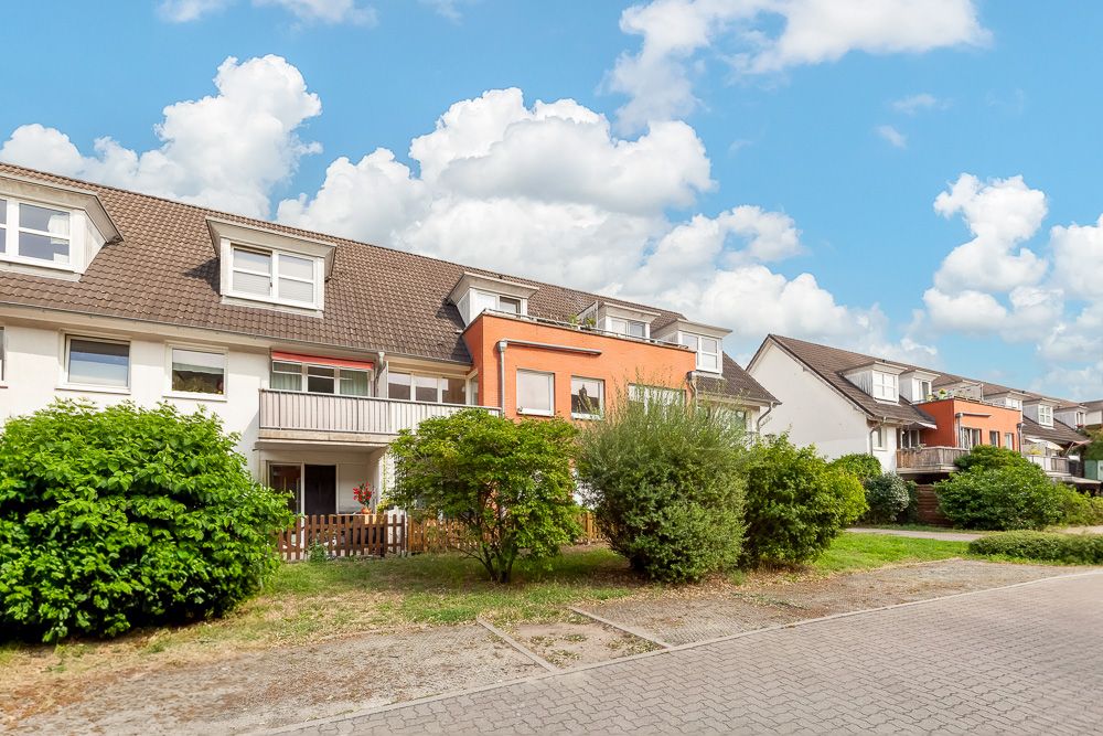 Reihenhaus mit Balkon, Vorgarten und Sträuchern an einer Wohnstraße bei blauem Himmel.