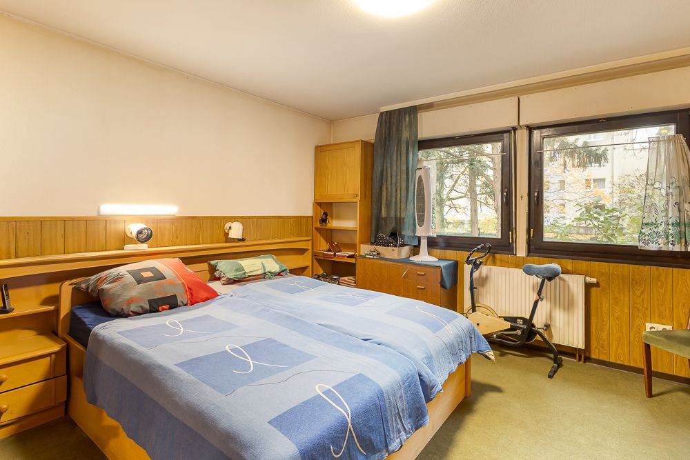 Schlafzimmer mit Doppelbett, Nachttischen, Heimtrainer und großen Fenstern mit Vorhängen.