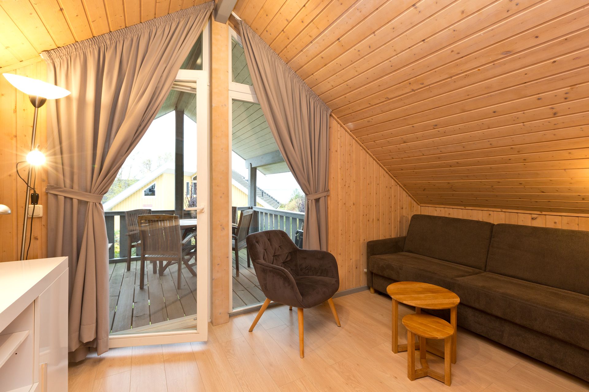 Wohnzimmer mit Holzdecke, braunem Sofa, Sessel, Couchtischen und Zugang zum Balkon.
