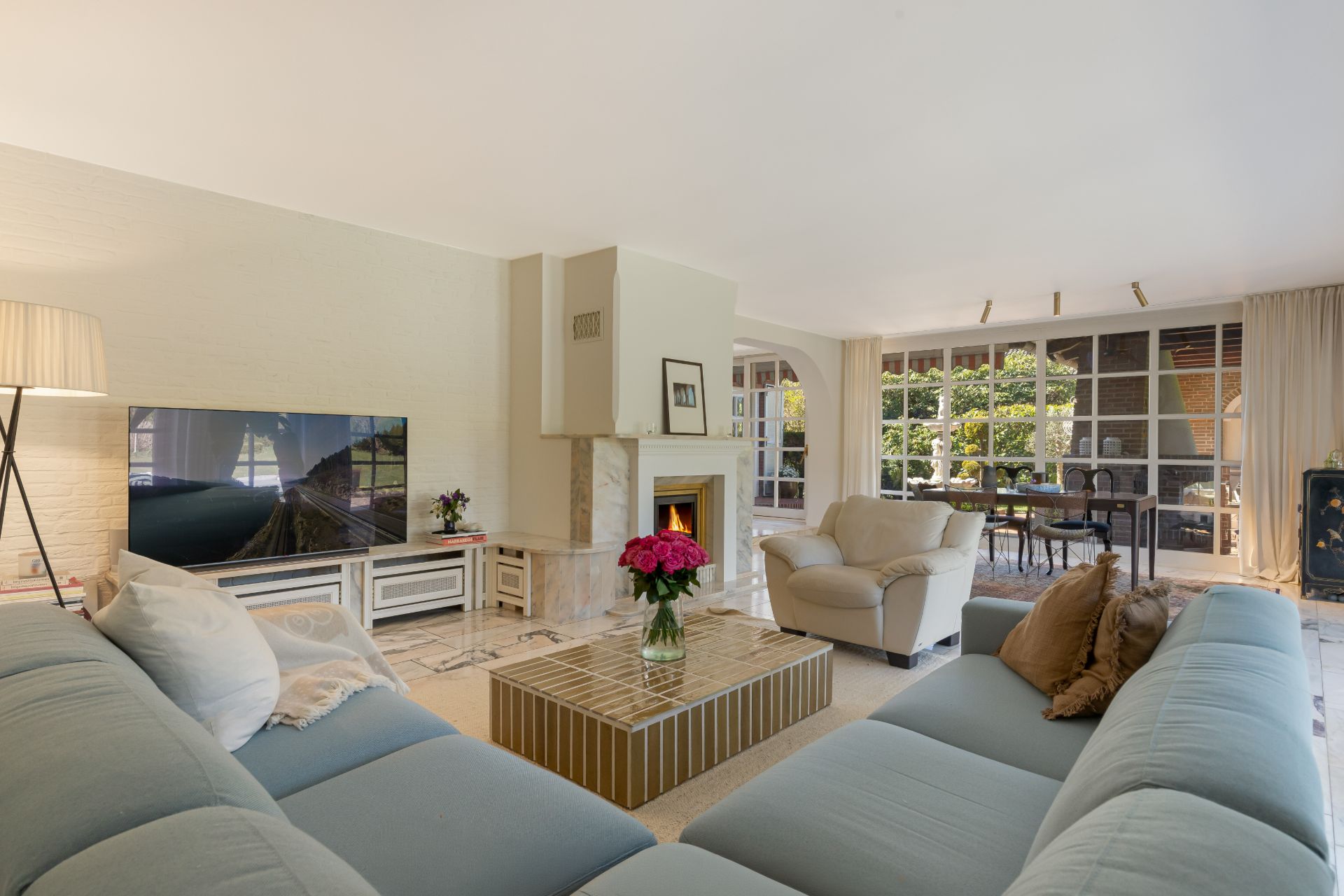 Helles Wohnzimmer mit großem Sofa, Couchtisch, Fernseher, Kamin und bodentiefen Fenstern.