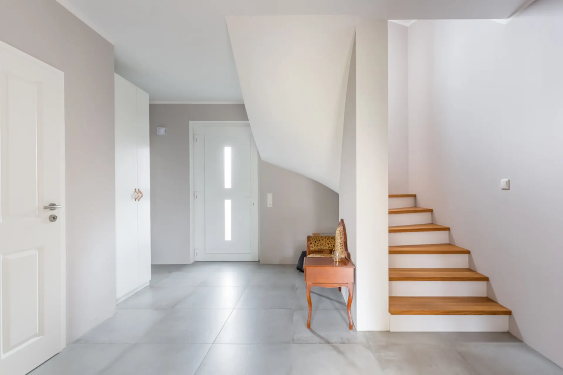 Heller Flur mit Treppe, weißer Tür, Schrank und Holzbank unter der Treppe.