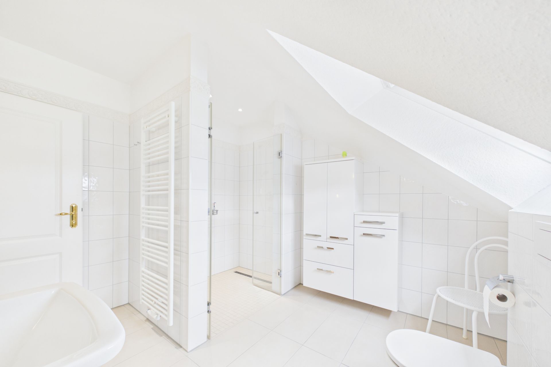 Helles Badezimmer mit Badewanne, Dusche, weißen Fliesen und Schränken unter einer Dachschräge.