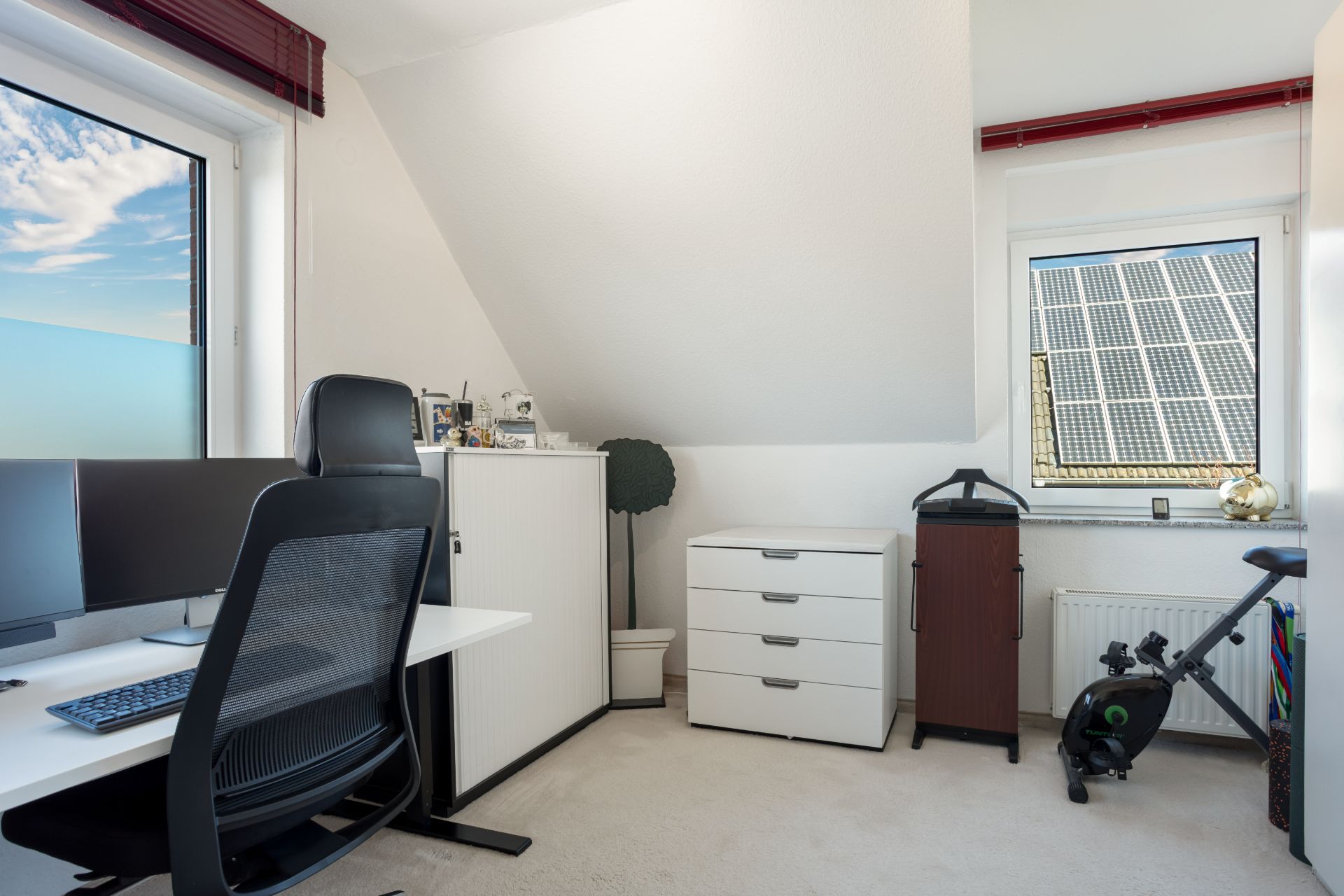 Helles Arbeitszimmer mit Schreibtisch, Stuhl, Monitoren, Kommode und Heimtrainer unter Dachschräge.