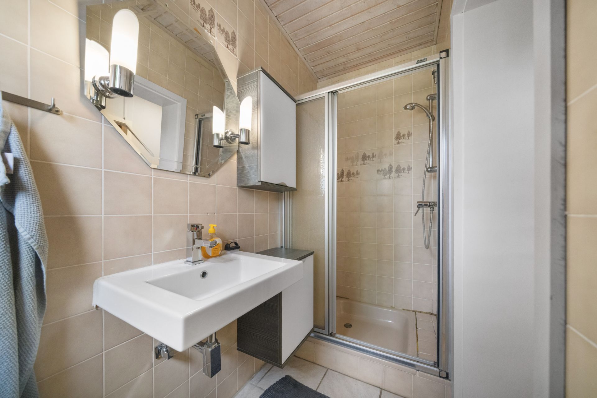 Kleines Badezimmer mit Waschbecken, Spiegel, Duschkabine und beiger Fliesenwand.