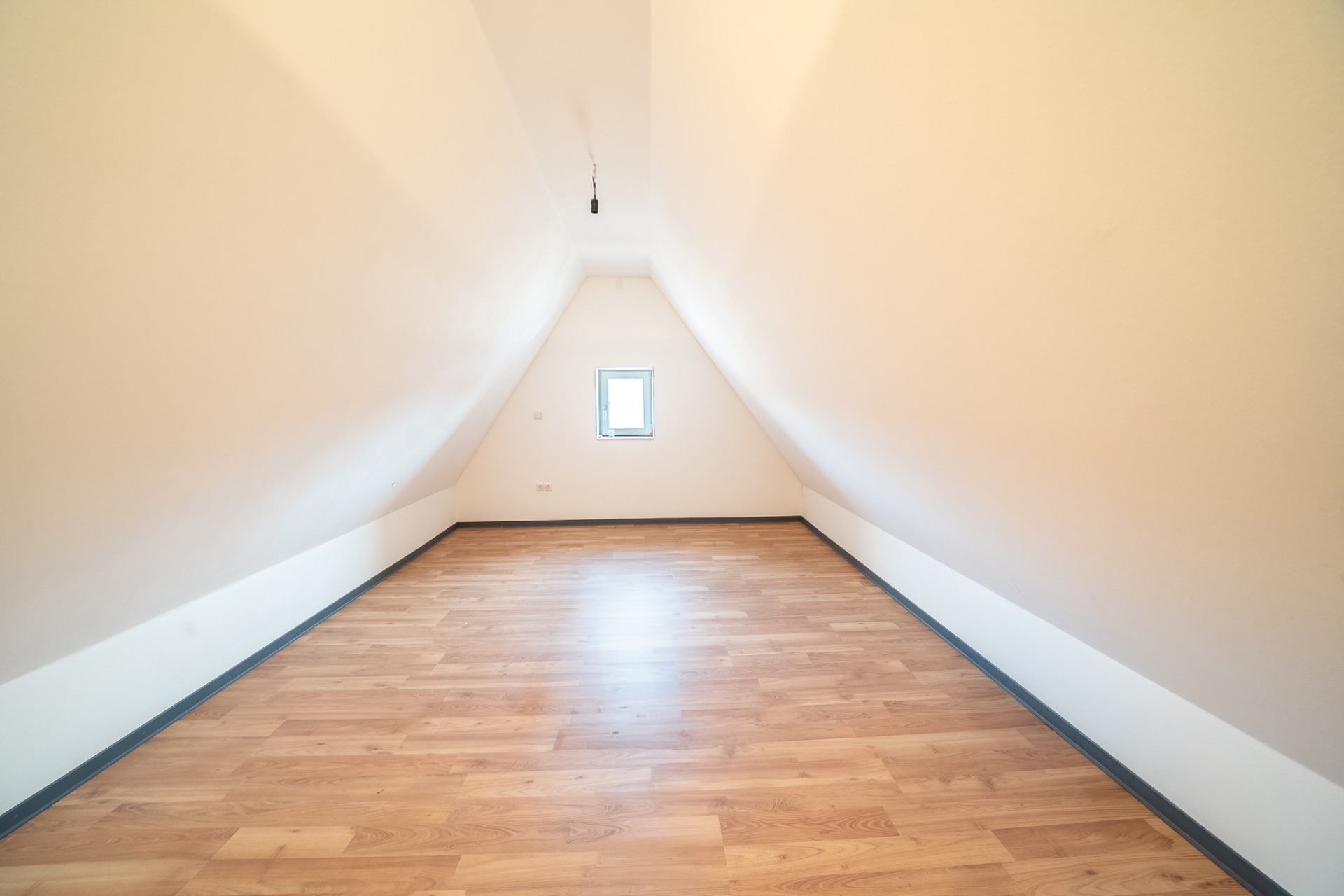 Leerer, heller Dachbodenraum mit schrägen Wänden, Holzboden und kleinem Fenster an der Stirnseite.