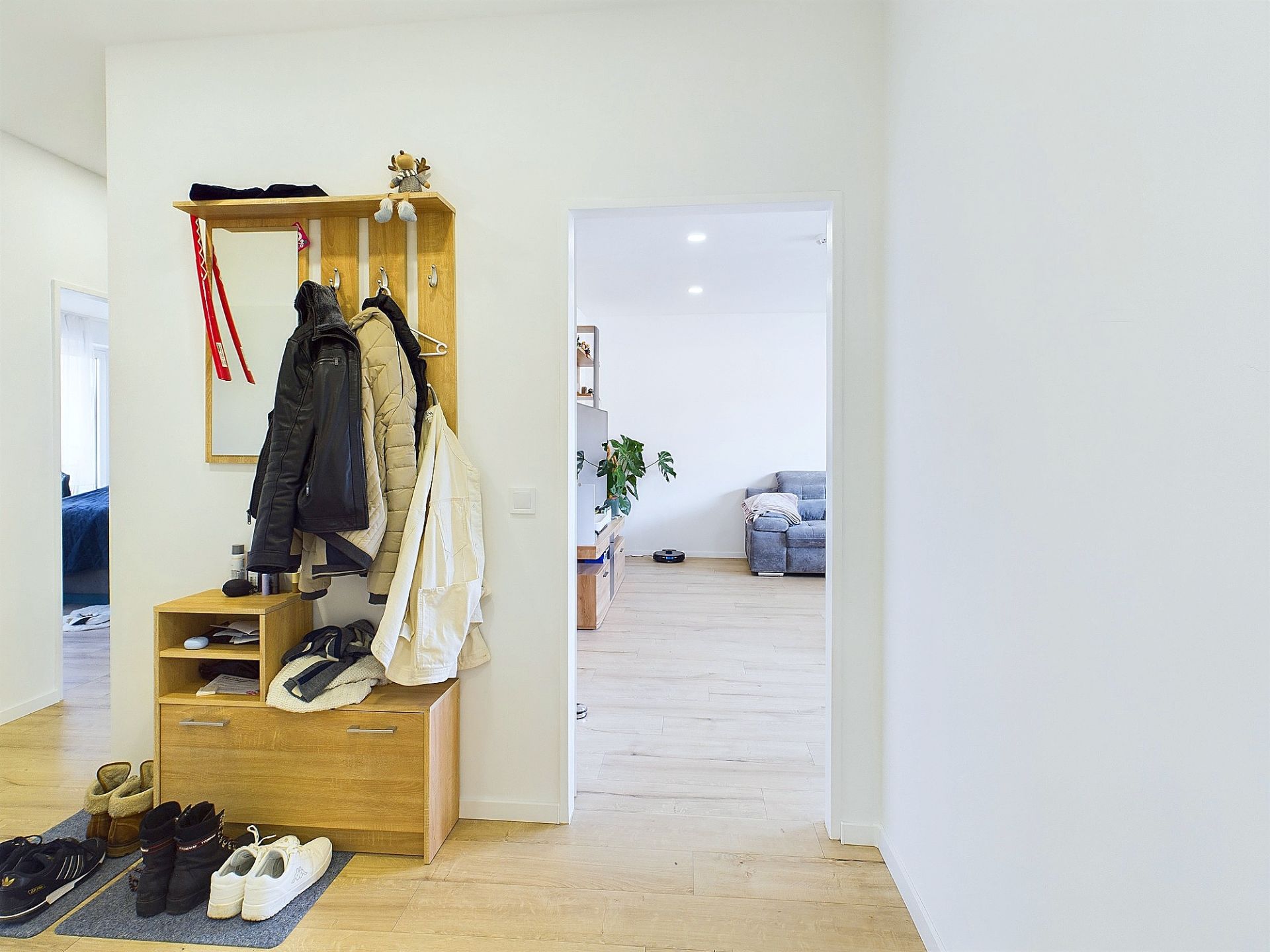 Flur mit Garderobe, Jacken, Schuhen und Blick ins Wohnzimmer mit Sofa und Pflanze.
