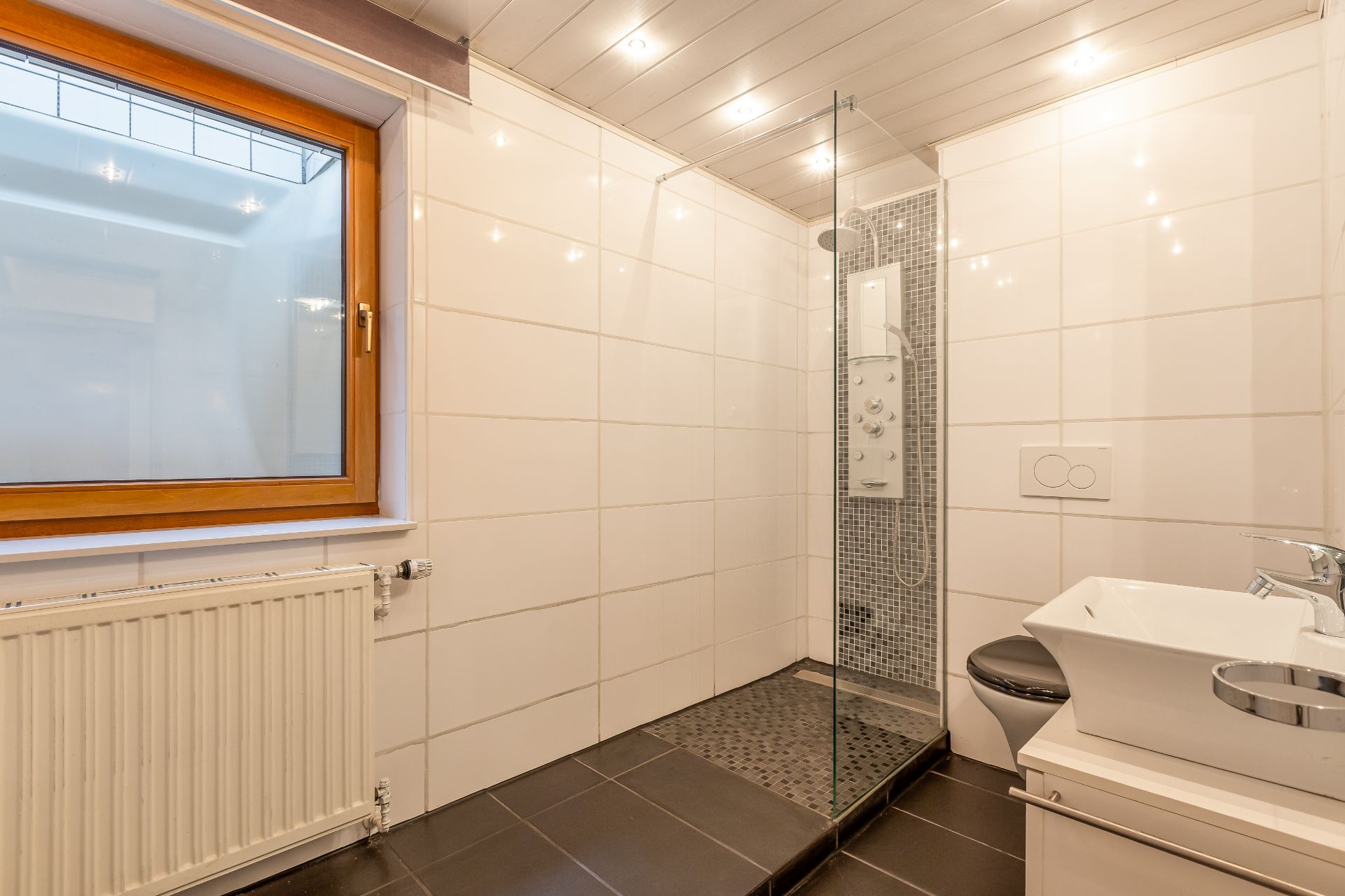 Modernes Badezimmer mit großer Dusche, Waschbecken, WC, Fenster und Fliesenboden.