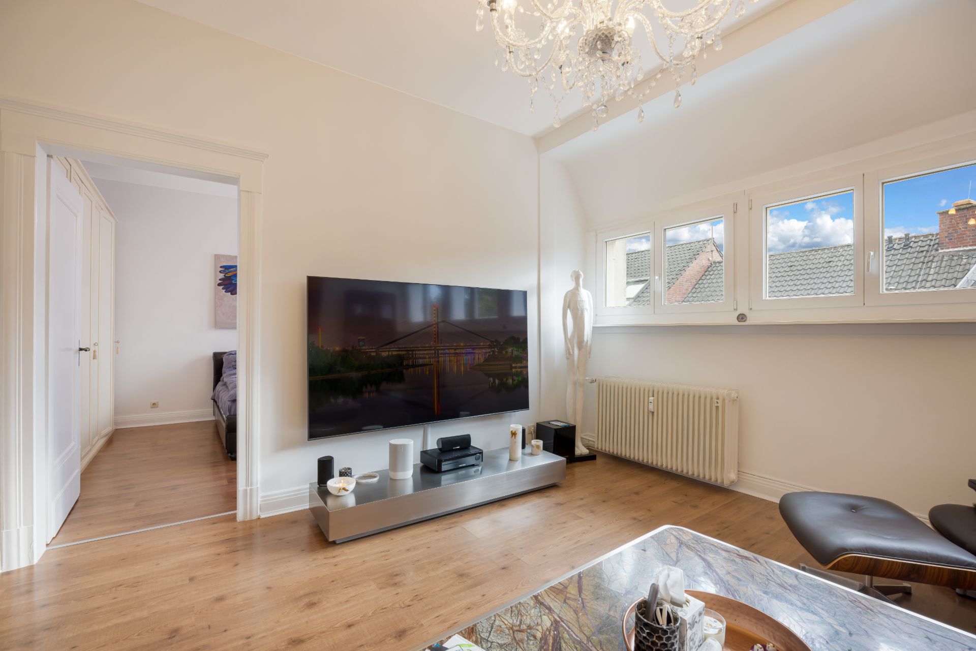 Wohnzimmer mit Fernseher, Sessel, Flachheizkörper und Fenstern mit Blick aufs Dach.