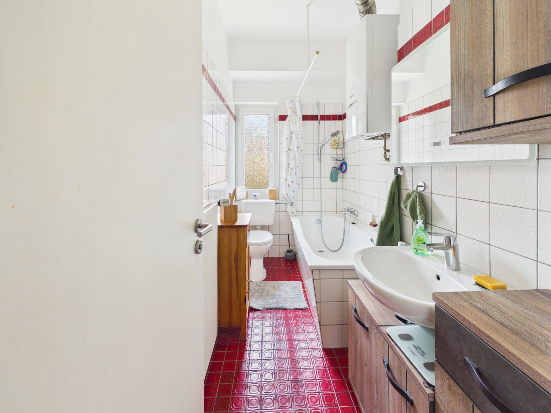Helles Bad mit rotem Fliesenboden, Badewanne, Waschbecken, Toilette und Holzschränken.