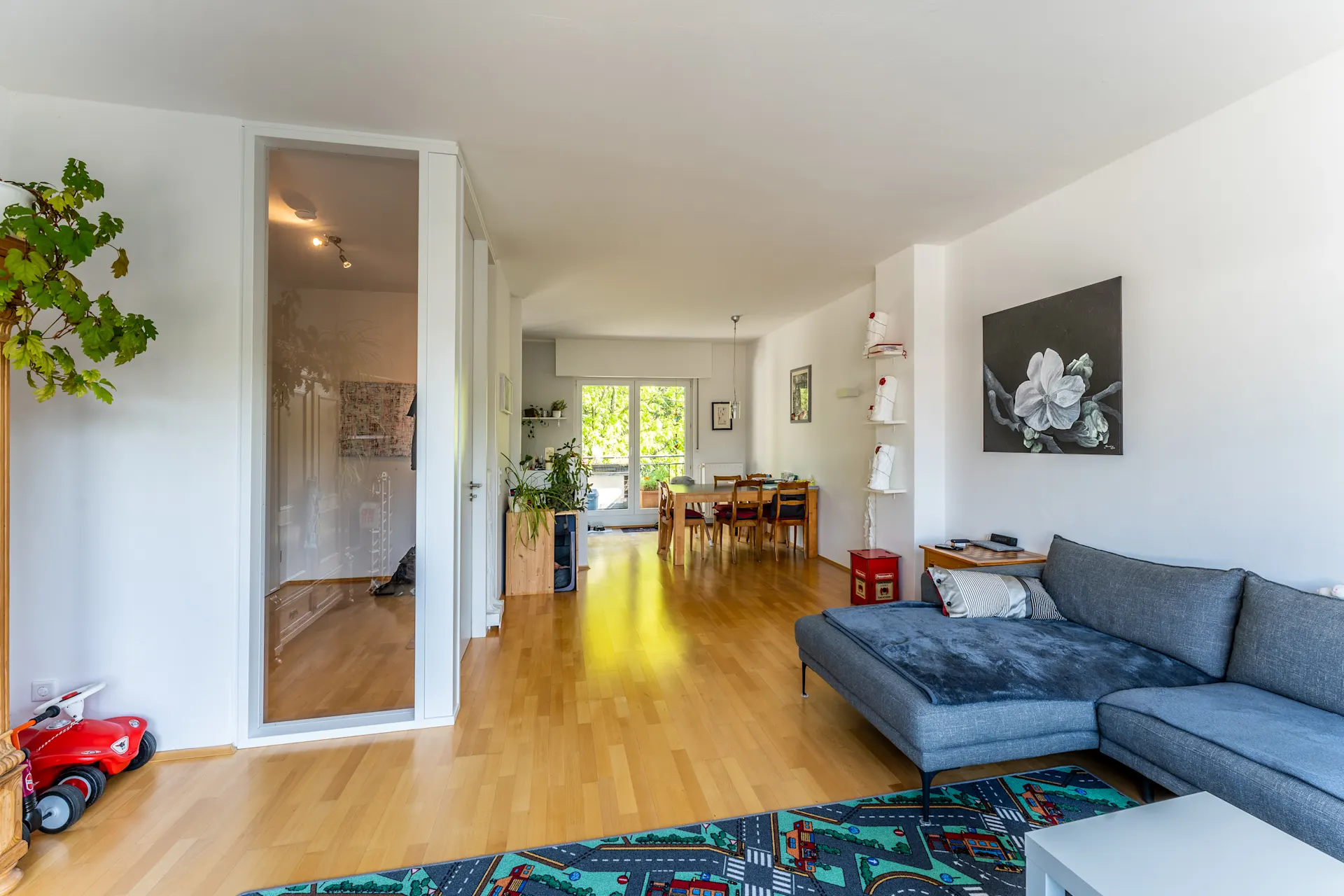 Helles Wohnzimmer mit grauem Sofa, Esstisch, Pflanzen und großem Fenster im Hintergrund.