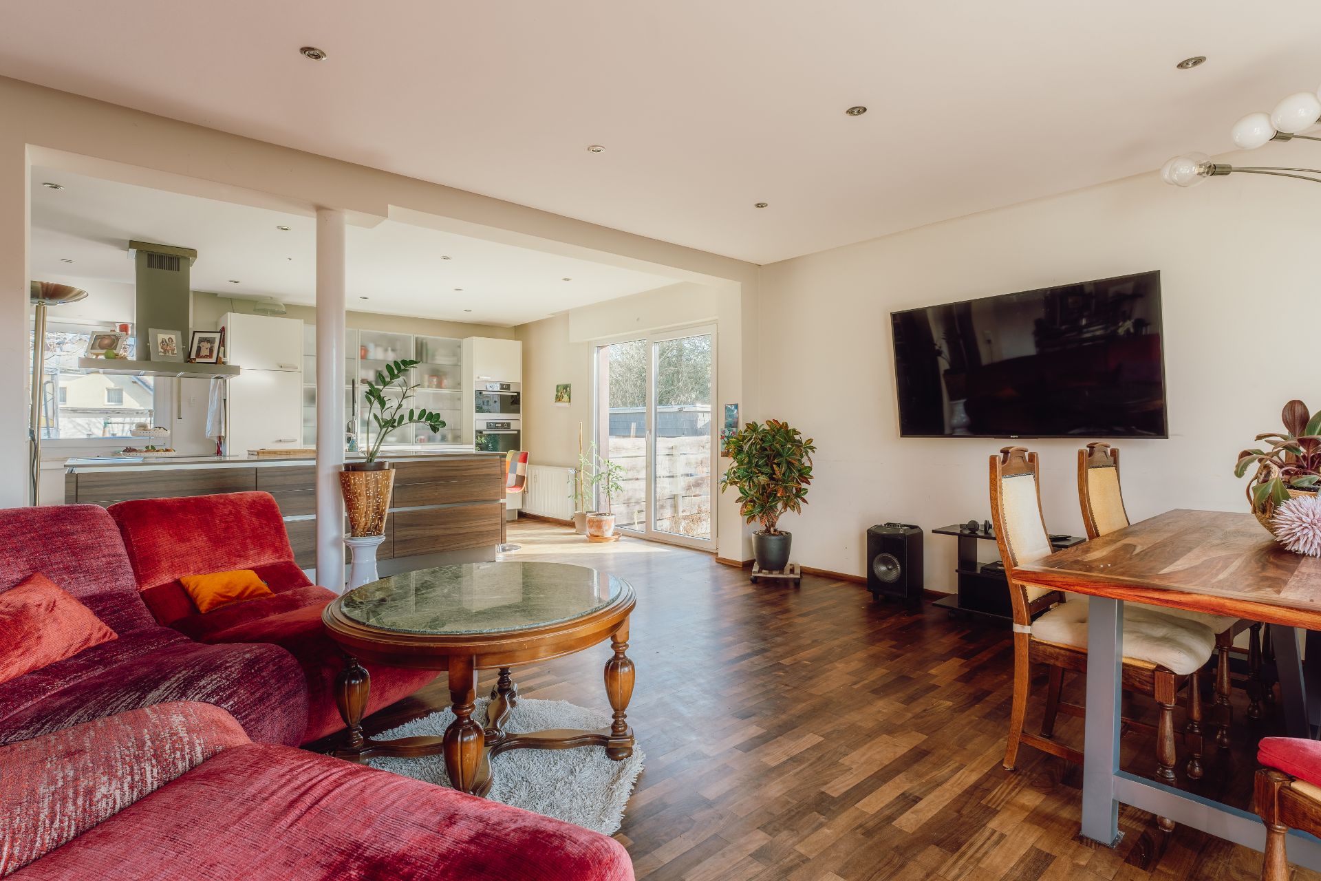 Wohnzimmer mit rotem Sofa, Esstisch, TV an der Wand und offener Küche im Hintergrund.