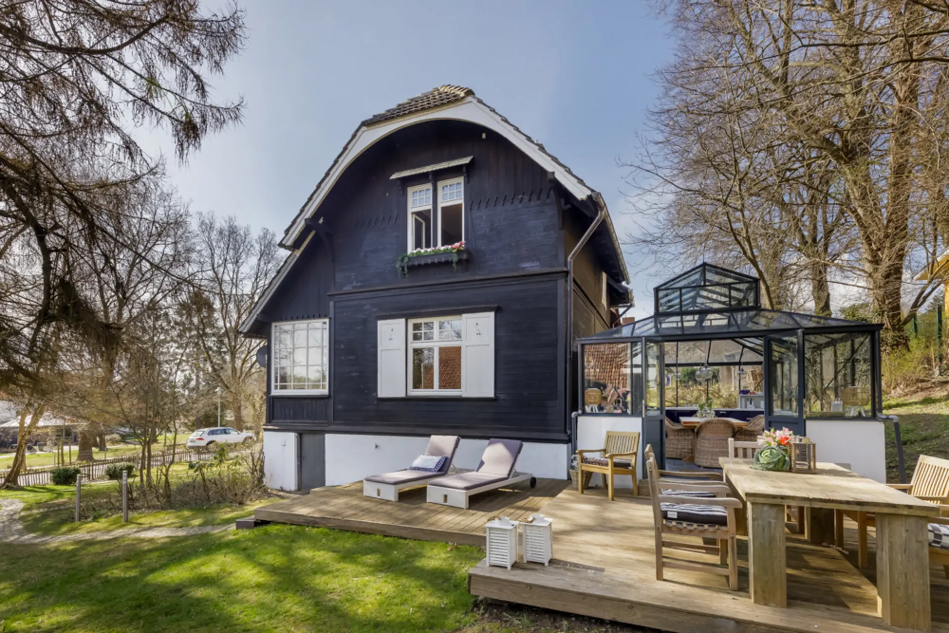 Holzhaus mit Terrasse, Wintergarten und Gartenmöbeln, umgeben von Bäumen.