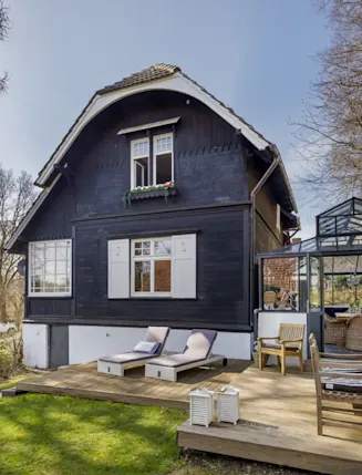 Holzhaus mit Terrasse, Wintergarten und Gartenmöbeln, umgeben von Bäumen.