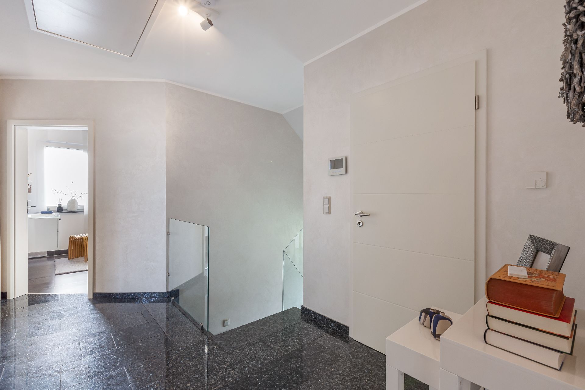 Heller Flur mit schwarzem Boden, weißer Tür und Treppe mit Glasgeländer.
