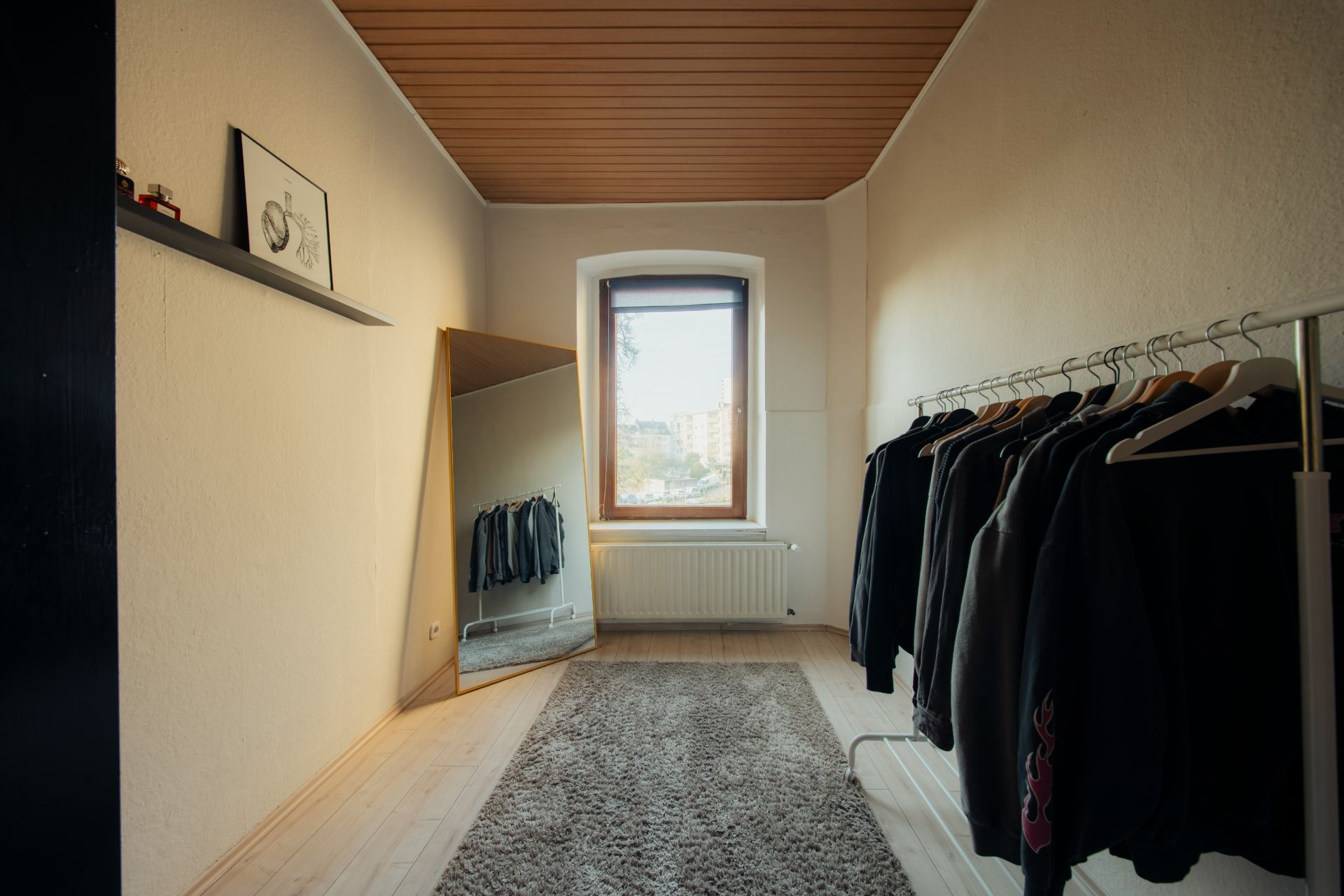 Zimmer mit Teppich, großem Spiegel, Fenster und Kleiderstange mit dunkler Kleidung.