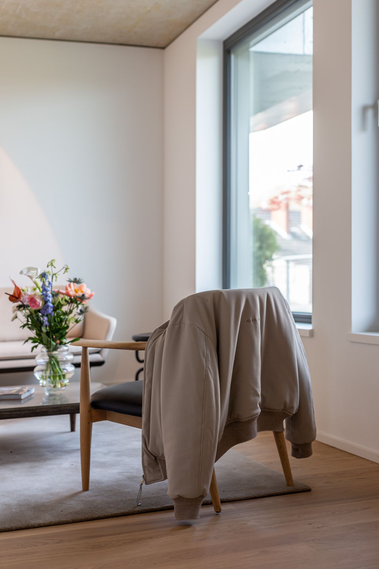 Stuhl mit beiger Jacke, daneben ein Glastisch mit Blumenstrauß in hellem Wohnzimmer.