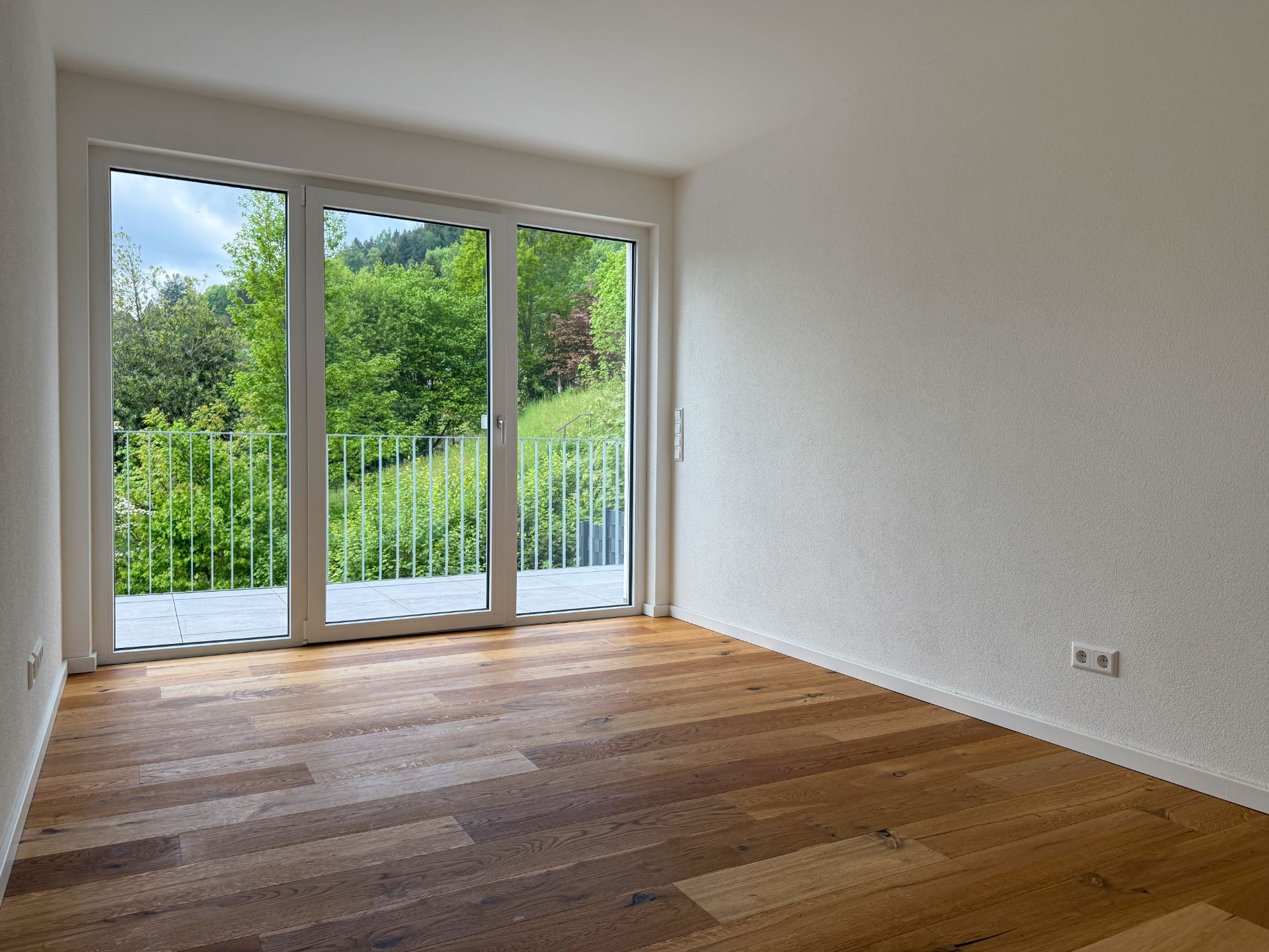 Leerer Raum mit Holzfußboden und großen Fenstern mit Blick ins Grüne.