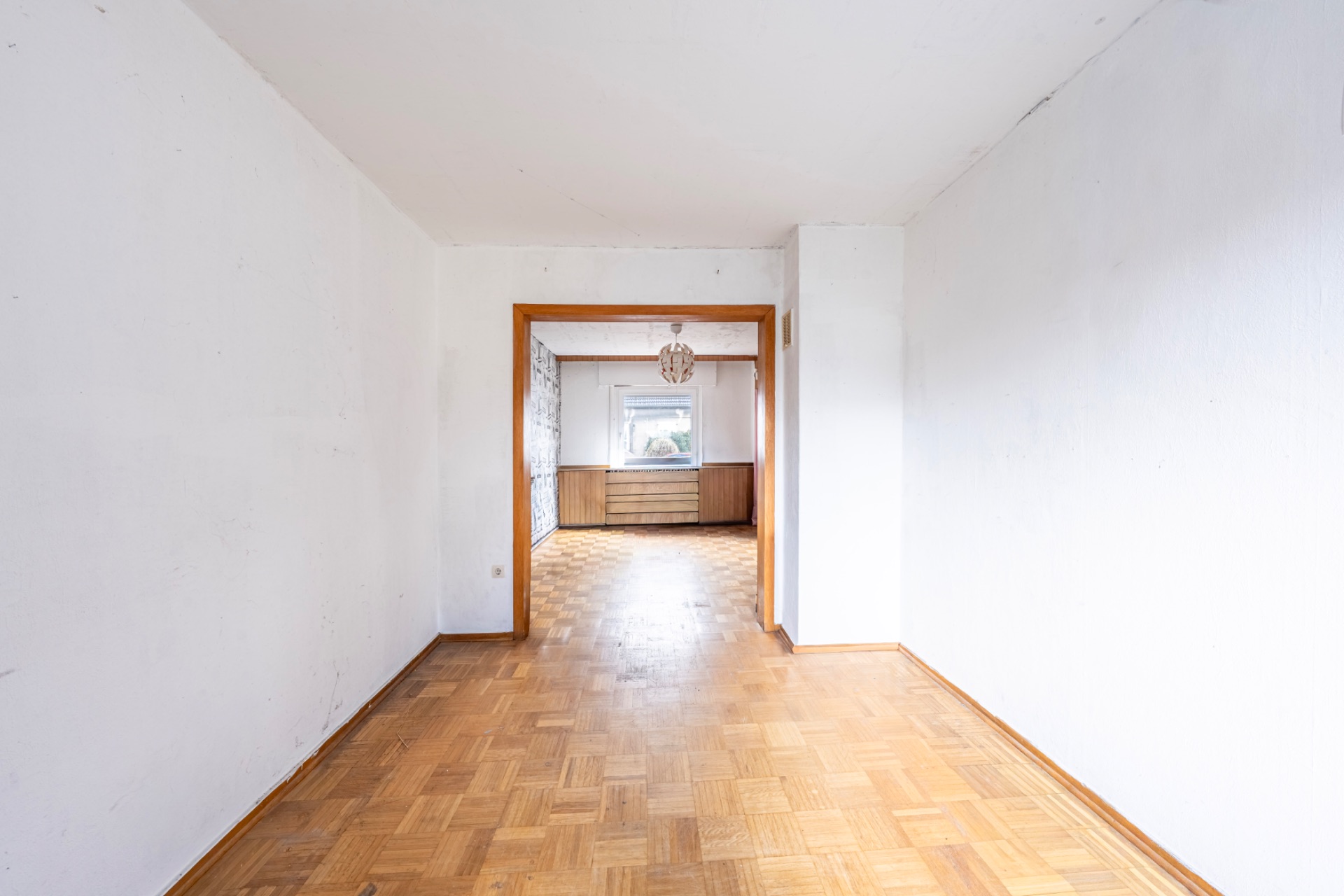 Leerer Raum mit Parkettboden, offenem Durchgang und Fenster im Hintergrund.