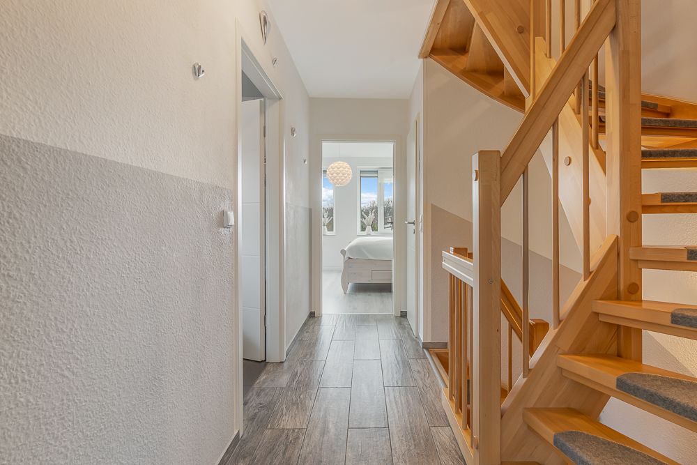 Heller Flur mit Holztreppe und Blick in ein Schlafzimmer mit Fenster.