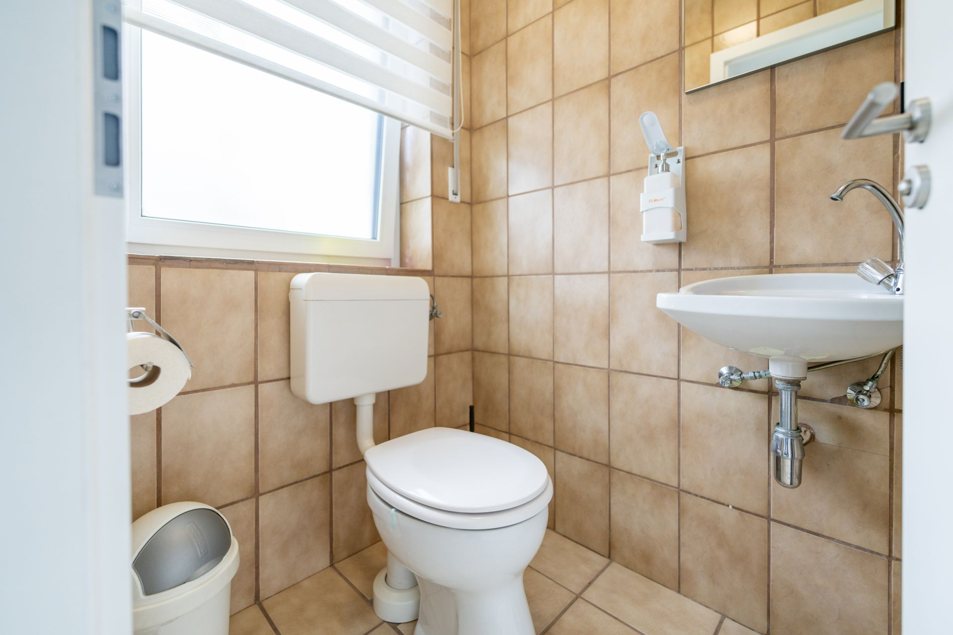 Kleines Badezimmer mit Toilette, Waschbecken, Spiegel und Fenster, Fliesen an Wand und Boden.