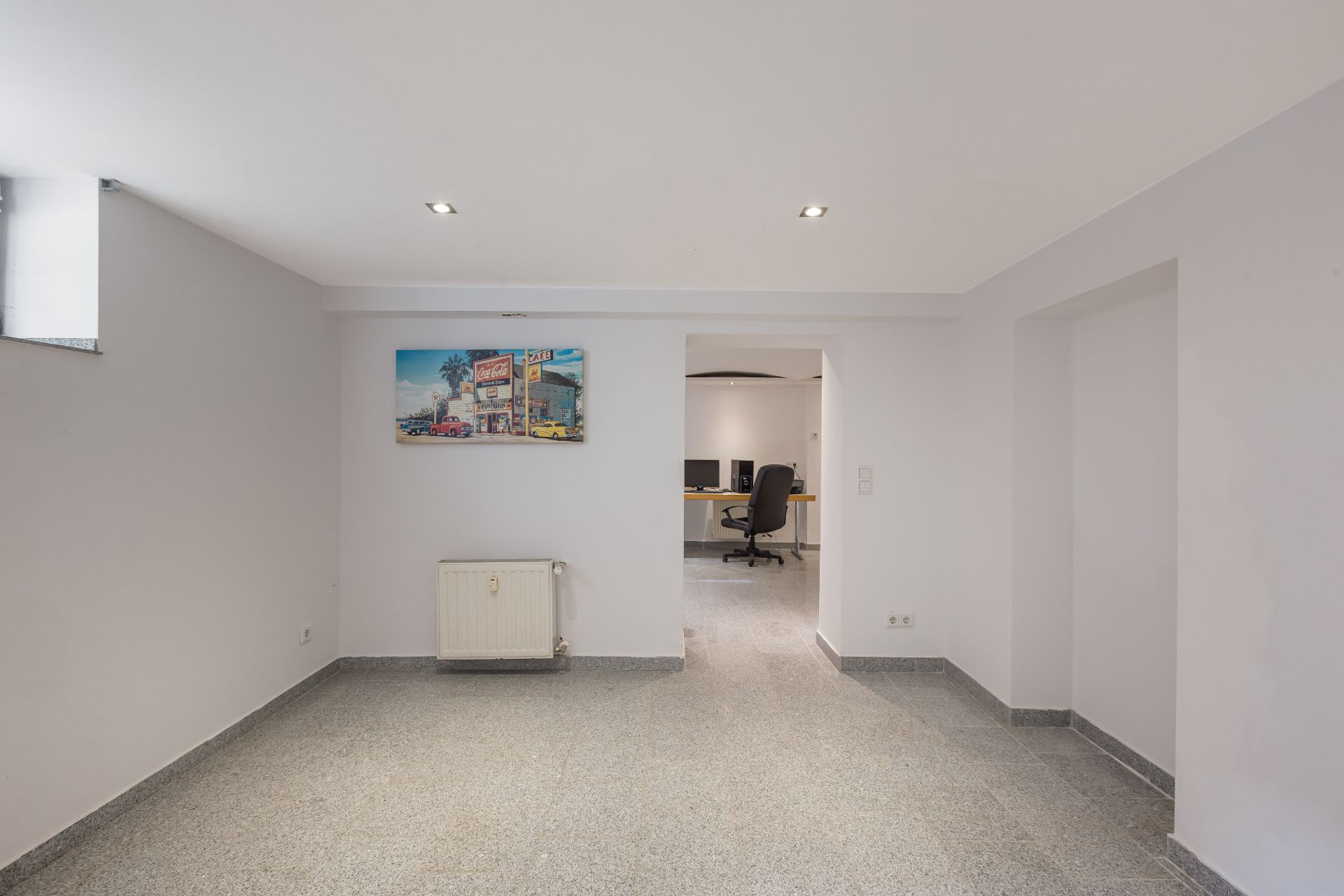 Heller Raum mit grauem Boden, Wandbild, Heizkörper und Blick auf Arbeitsbereich mit Schreibtisch und Stuhl.