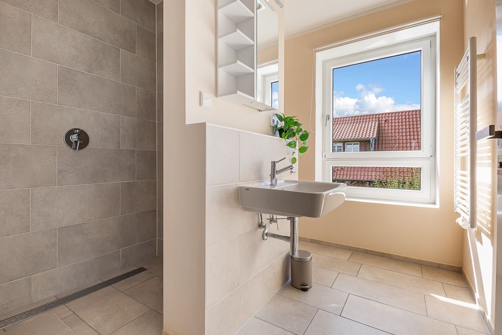 Helles Badezimmer mit Dusche, Waschbecken, großem Fenster und Handtuchheizkörper.