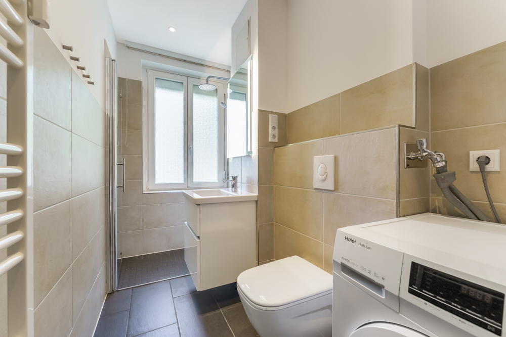 Modernes Bad mit Dusche, Waschbecken, WC und Waschmaschine, große Fenster lassen Tageslicht herein.