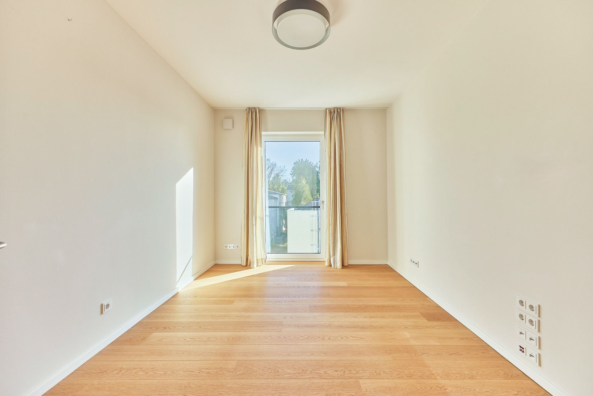 Helles, leeres Zimmer mit Holzfußboden und großem Fenster mit Vorhängen.