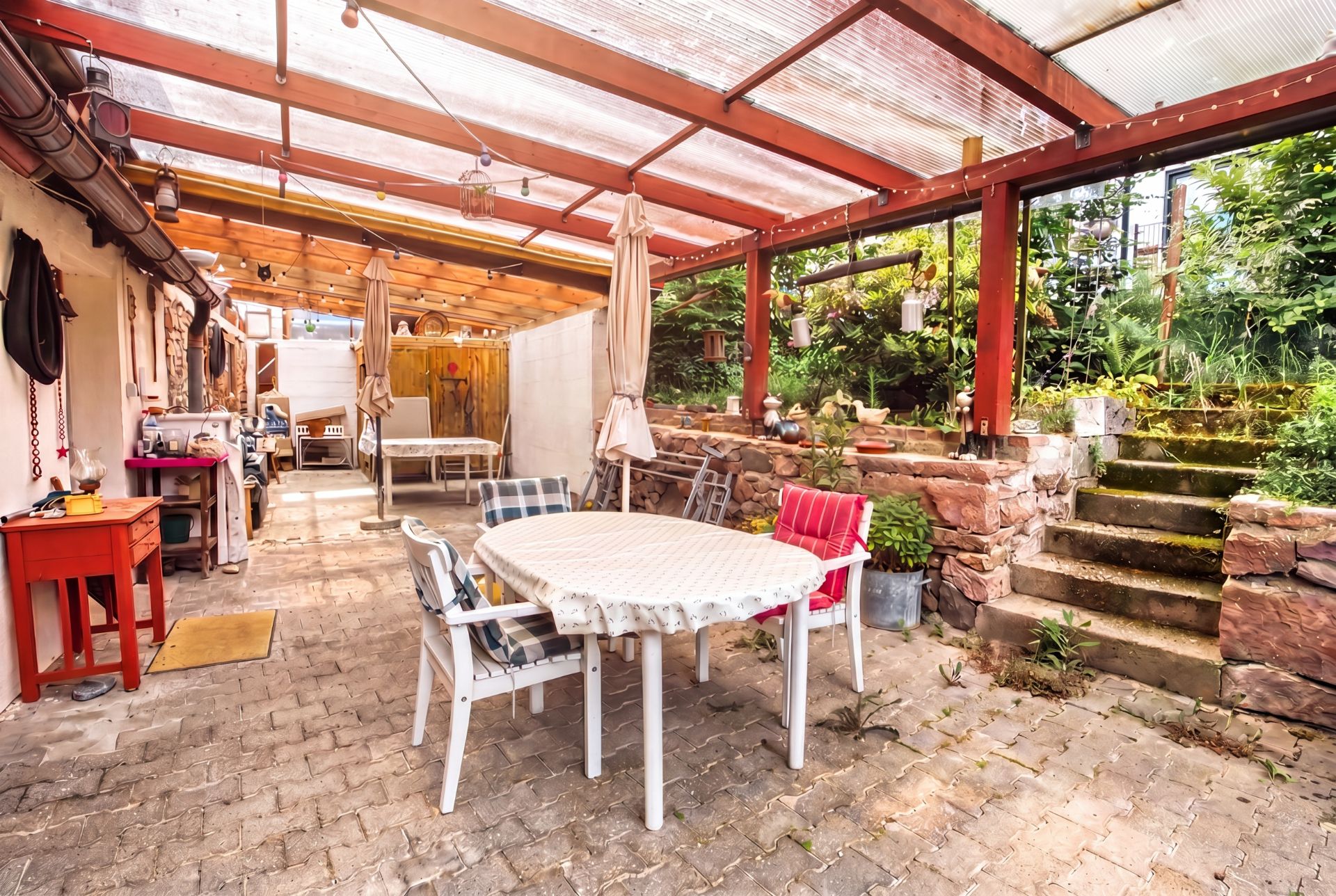 Überdachte Terrasse mit rundem Tisch und Stühlen, daneben eine Steintreppe zum Garten.