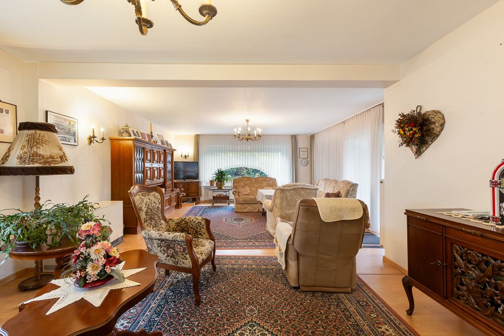 Helles Wohnzimmer mit Teppichen, Sesseln, Sofa, Schrankwand, Pflanzen und Kronleuchtern.