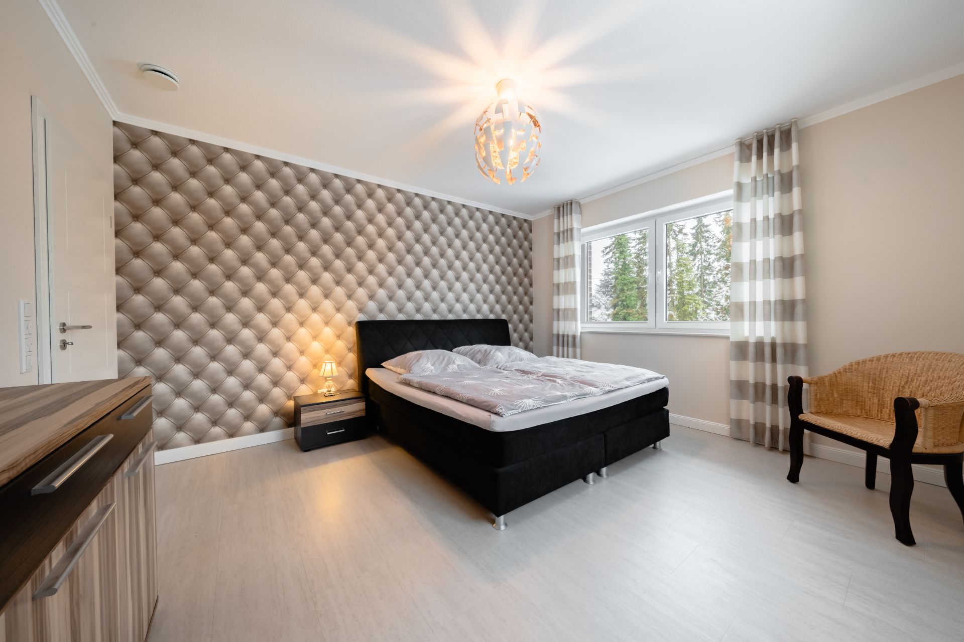 Helles Schlafzimmer mit Doppelbett, Nachttisch, Sessel und großem Fenster mit Vorhängen.