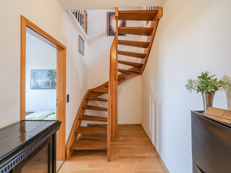 Heller Flur mit Holztreppe, Holzboden und offener Tür zu einem angrenzenden Zimmer.