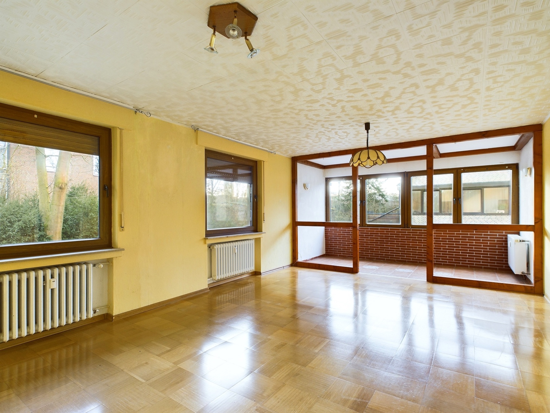 Leerer Wohnraum mit großen Fenstern, Parkettboden und Zugang zu einem Wintergarten.