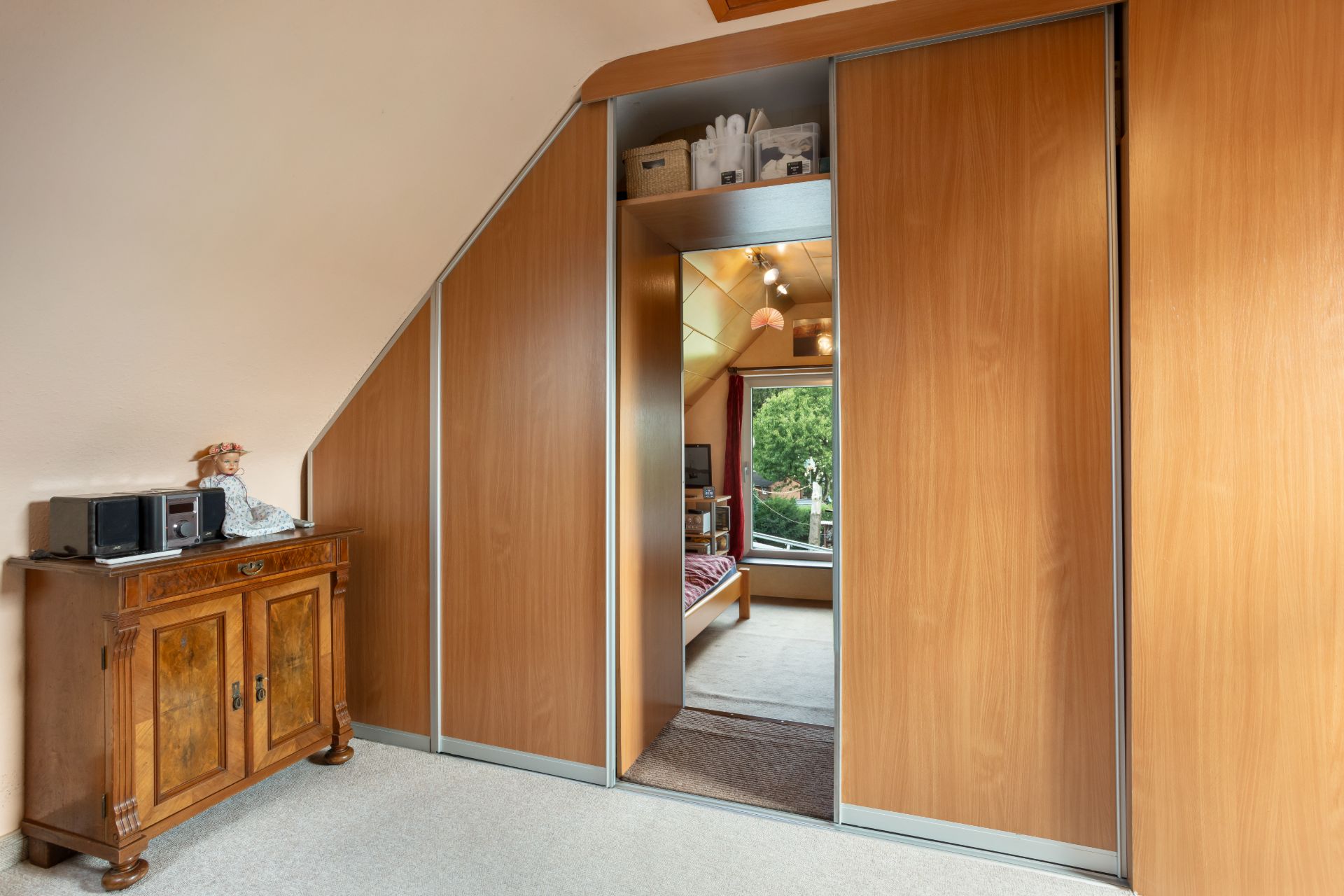 Holzschrank mit Schiebetüren unter Dachschräge, links steht eine Kommode mit Geräten.