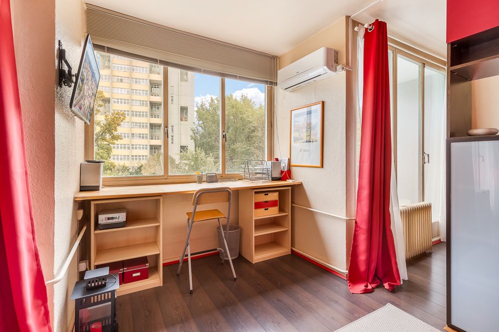 Kleines Arbeitszimmer mit Schreibtisch am Fenster, Stuhl, Regalen und rotem Vorhang.